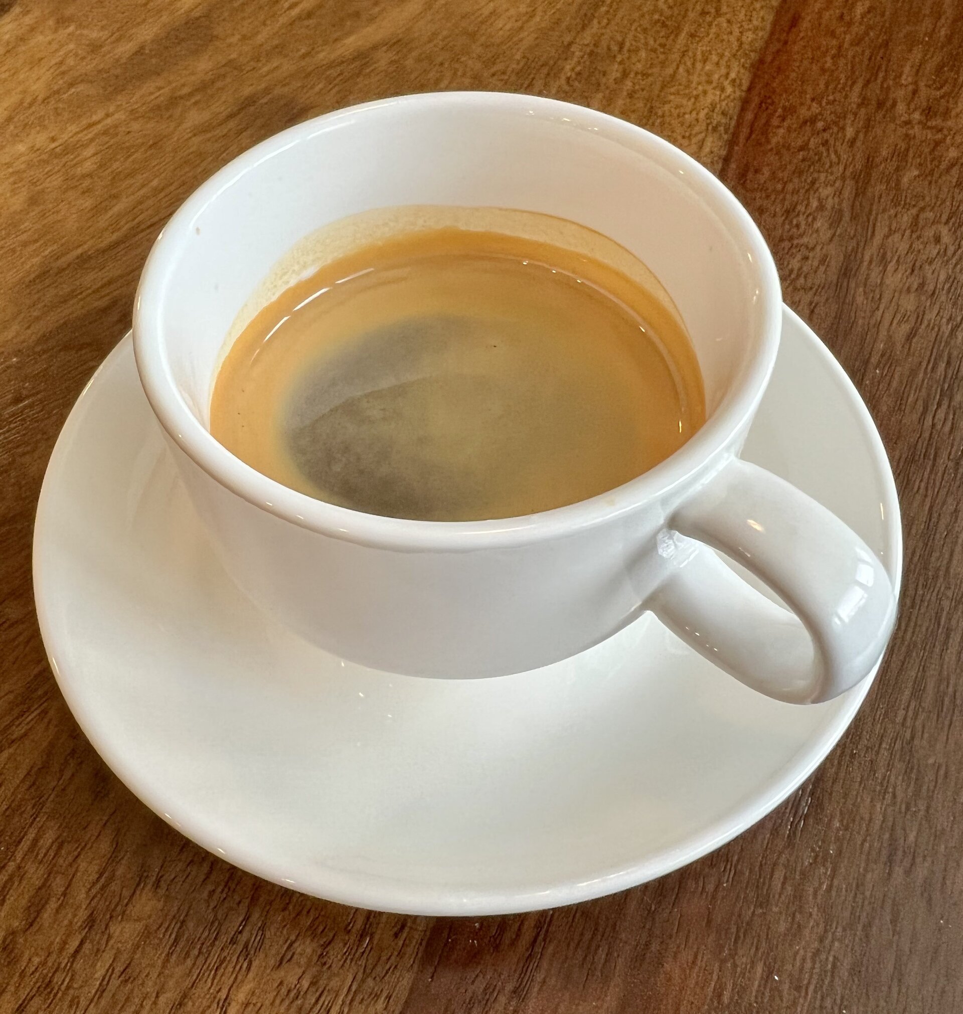 Un café expresso avec une crème marron clair dans une tasse à expresso blanche dotée d'une anse. La tasse est posée sur une soucoupe blanche sur une table en bois.