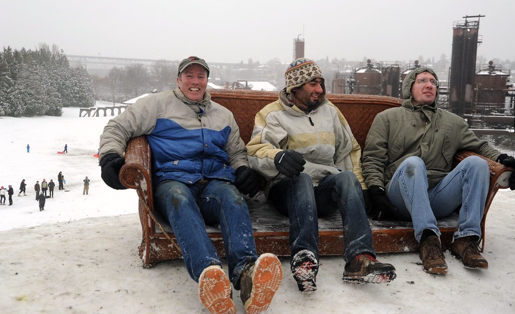 Trois hommes souriants sont assis sur un canapé en osier, sans coussins, au sommet d'une colline couverte de neige tassée. Ils portent des vestes d'hiver, des gants, des jeans mouillés et des chaussures recouvertes de neige. Une usine se profile en arrière‑plan et un groupe de personnes avec des luges est rassemblé en bas de la colline.