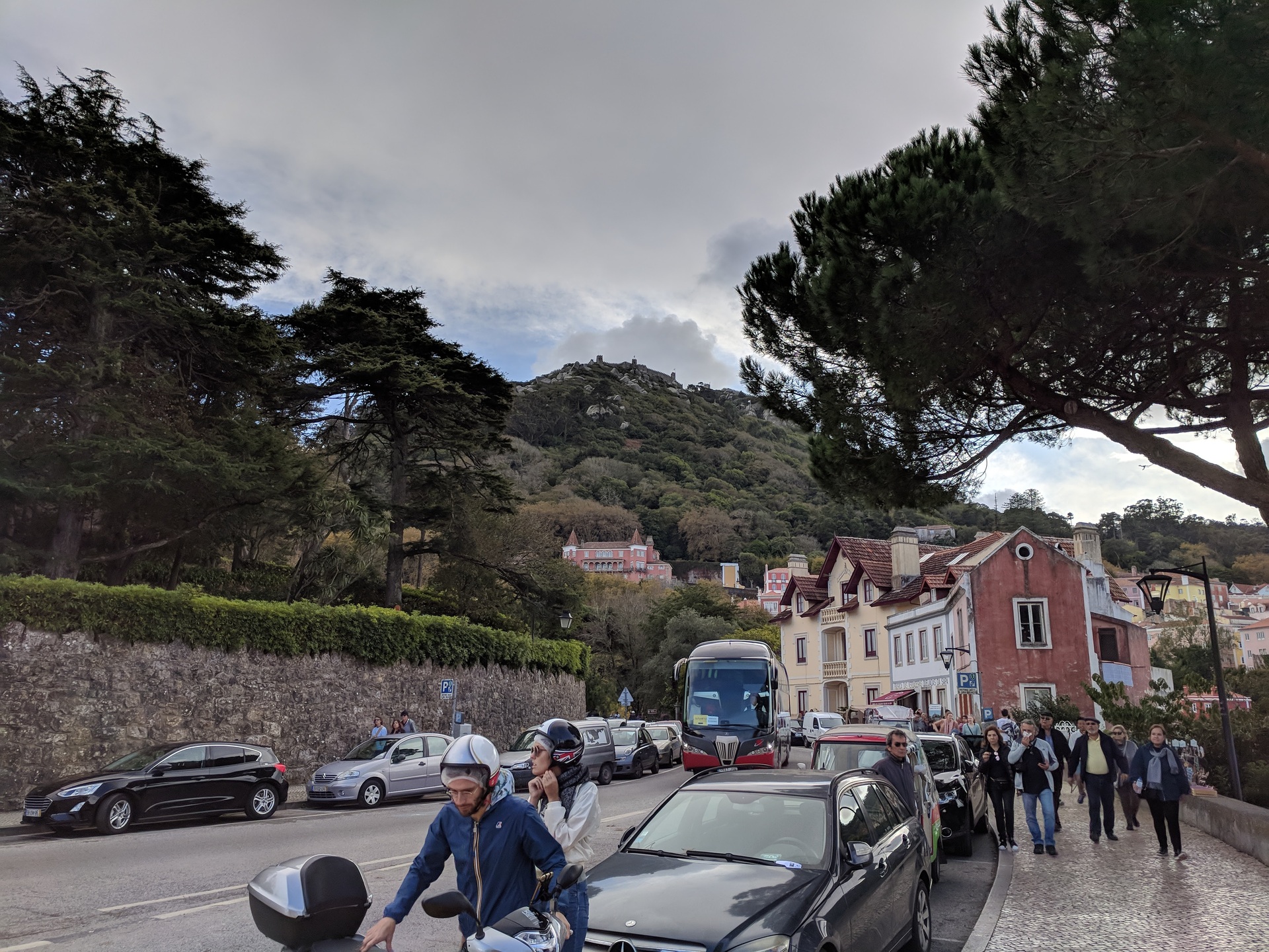 Vista de um trecho da cidade de Sintra, em Portugal, em um dia frio e nublado. Um ônibus de turismo desce a rua, um casal desmonta de uma scooter e um grupo de pessoas caminha em nossa direção. No topo do morro, à distância, vê-se Castelo dos Mouros.